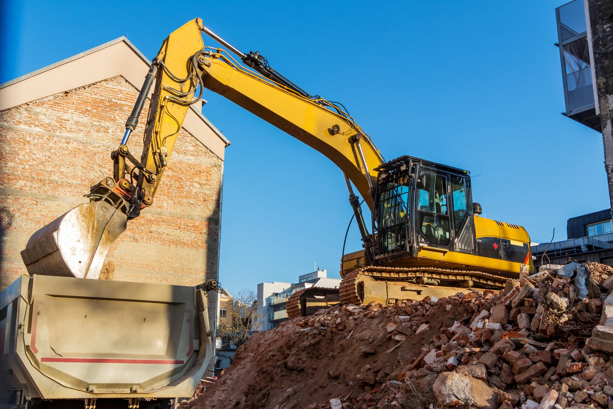 Excavator demolishing a brick house beside a skip bin